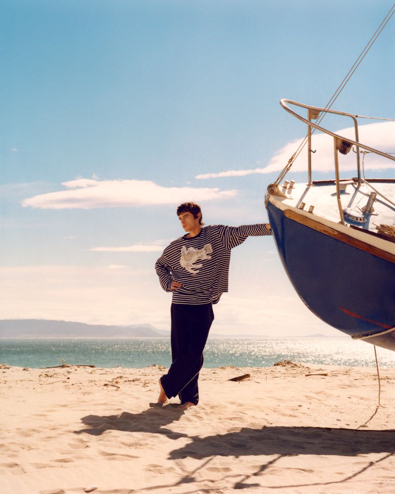 Man in a striped shirt stands barefoot on a sandy beach next to an upturned sailboat under a blue sky with wispy clouds.