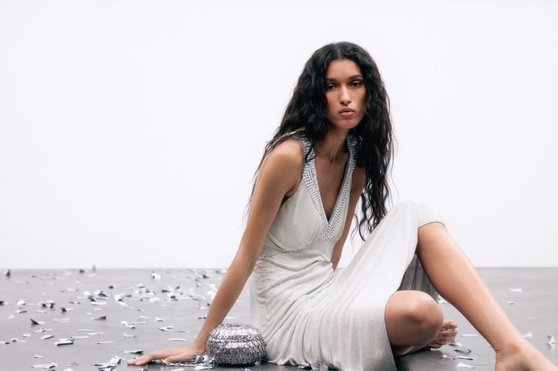 A slender woman with wavy hair poses gracefully on a gray surface amid silver confetti, wearing a sleeveless silver sequined dress in a studio.