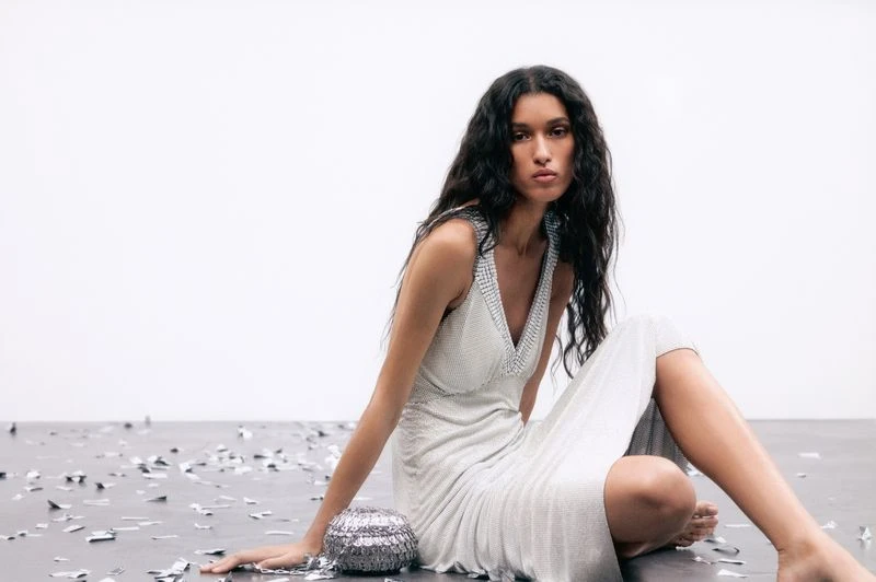 A slender woman with wavy hair poses gracefully on a gray surface amid silver confetti, wearing a sleeveless silver sequined dress in a studio.