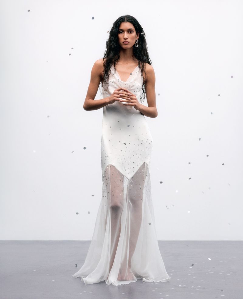 Elegant woman with long dark hair poses in a white embellished gown in a minimalist studio setting with confetti-like details scattered around.