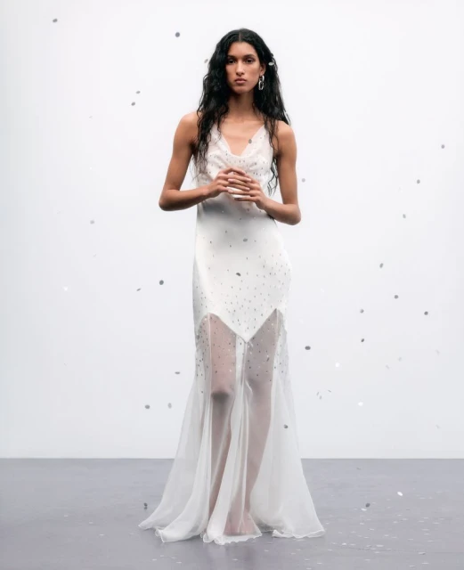 Elegant woman with long dark hair poses in a white embellished gown in a minimalist studio setting with confetti-like details scattered around.