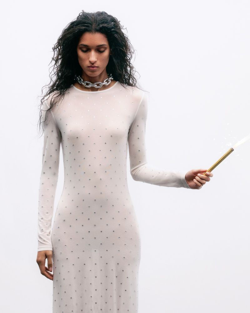 Model wearing a sheer studded long-sleeve dress and chunky silver chain necklace against a white background.