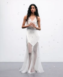 Model in white sheer dress standing against a light background with floating metallic confetti pieces
