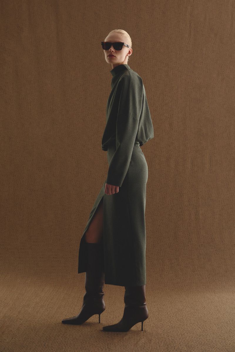 Full-length fashion portrait of a woman with a shaved head wearing a dark green dress, black sunglasses, and knee-high stiletto boots.