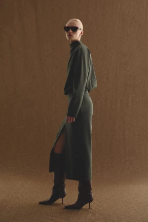 Full-length fashion portrait of a woman with a shaved head wearing a dark green dress, black sunglasses, and knee-high stiletto boots.