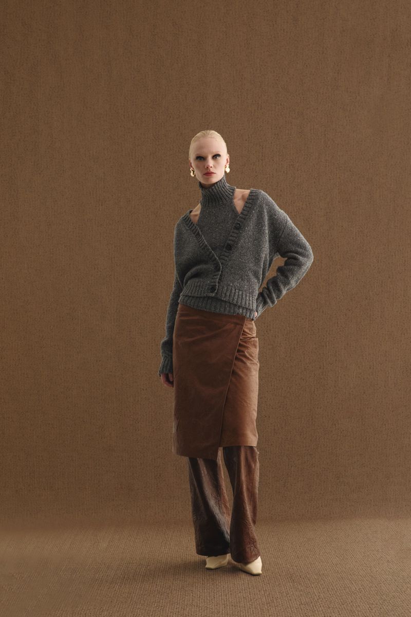 A woman poses in a grey cardigan, brown skirt over pants, and high heels against a textured brown backdrop, showcasing a fashion-forward look.