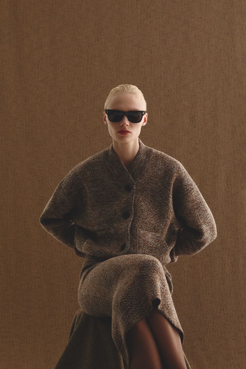 Blonde woman in sunglasses poses in a tweed cardigan and midi skirt against a neutral brown background, fashion studio shot.