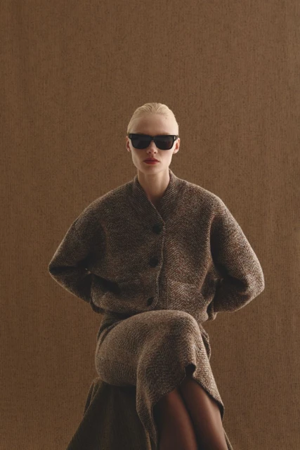 Blonde woman in sunglasses poses in a tweed cardigan and midi skirt against a neutral brown background, fashion studio shot.