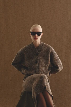 Blonde woman in sunglasses poses in a tweed cardigan and midi skirt against a neutral brown background, fashion studio shot.