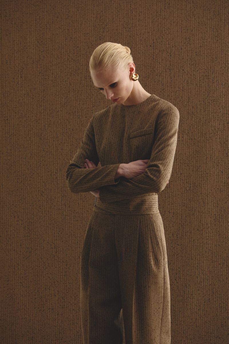 Fashion shot of a woman with her arms crossed. She wears a tan suit and gold earrings, against a textured tan background in studio light.