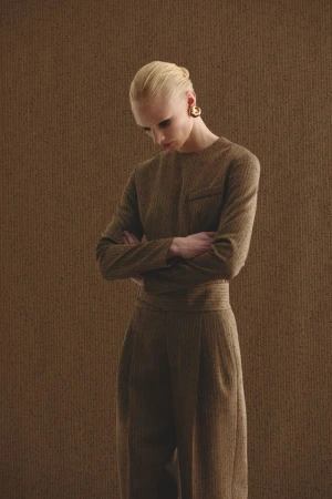 Fashion shot of a woman with her arms crossed. She wears a tan suit and gold earrings, against a textured tan background in studio light.