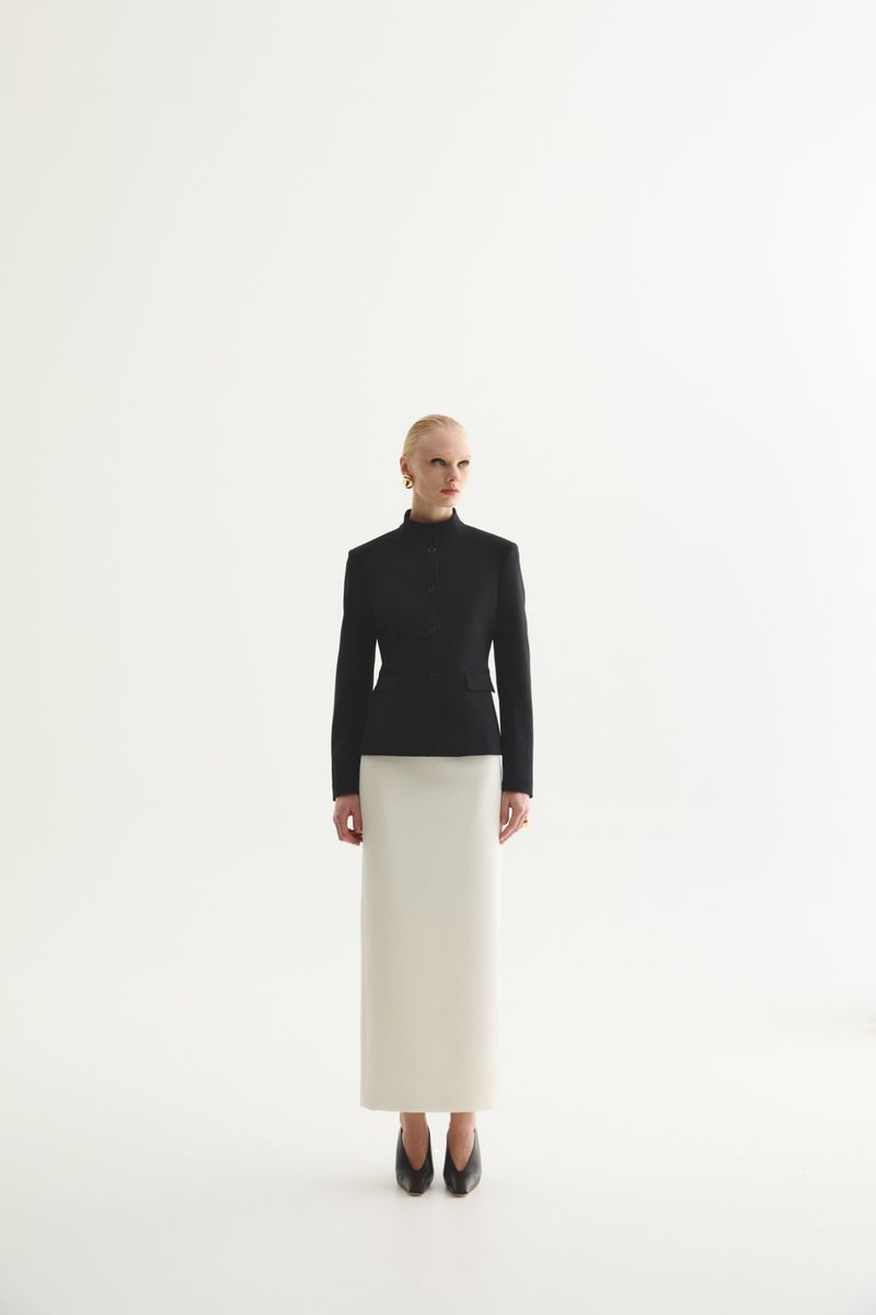 A fair-skinned adult female stands in a black jacket and cream skirt against a white background, radiating a sophisticated fashion editorial vibe.