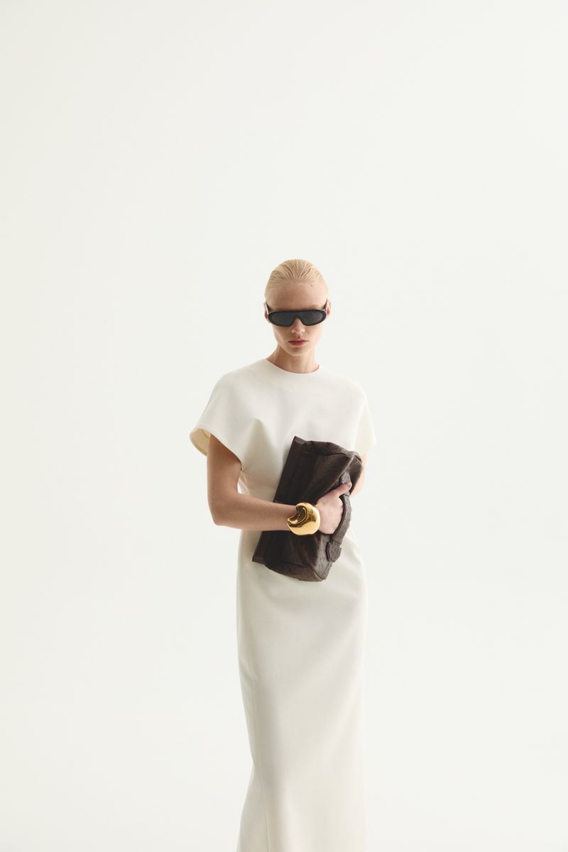 Woman posing in white dress and sunglasses holding a dark clutch with a gold bracelet, standing against a bright white backdrop in studio.
