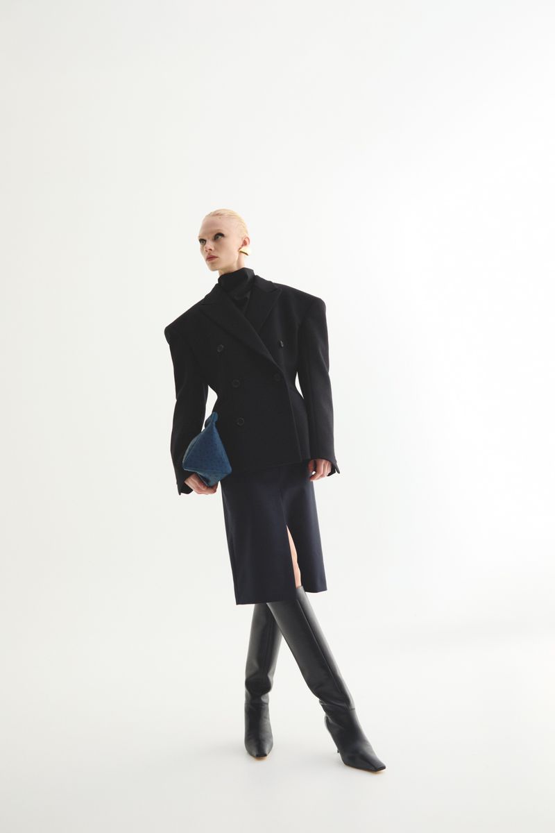 Fashionable adult female in a blazer dress and boots holding a clutch, posing in a photo studio with a neutral background.
