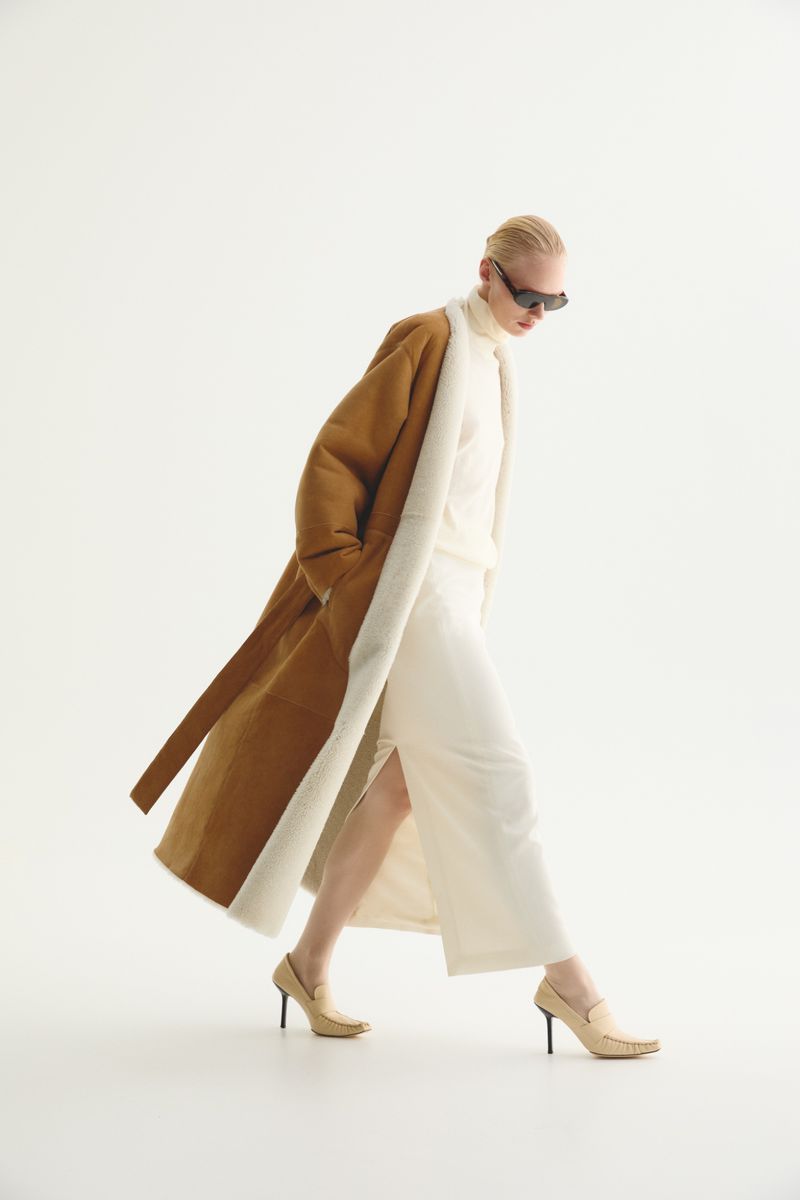 Fashion model wearing a tan coat and white clothing in a studio. She is wearing sunglasses and walking away from the camera.