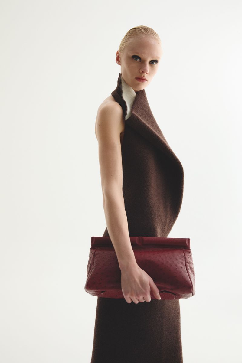 Fashion model posing in brown dress holding a red handbag against a white backdrop in a studio setting with neutral color palette.