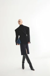 Model wearing a black structured blazer and tall boots holding a blue clutch against a white seamless background