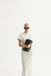 Model wearing a white dress, dark sunglasses, and holding a brown clutch against a clean white background