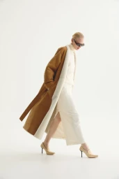 Model in sunglasses and a brown belted coat over a white outfit against a white background