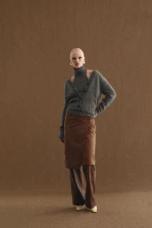 Model with a shaved head wearing a grey cardigan and brown skirt standing against a textured brown backdrop for Armarium