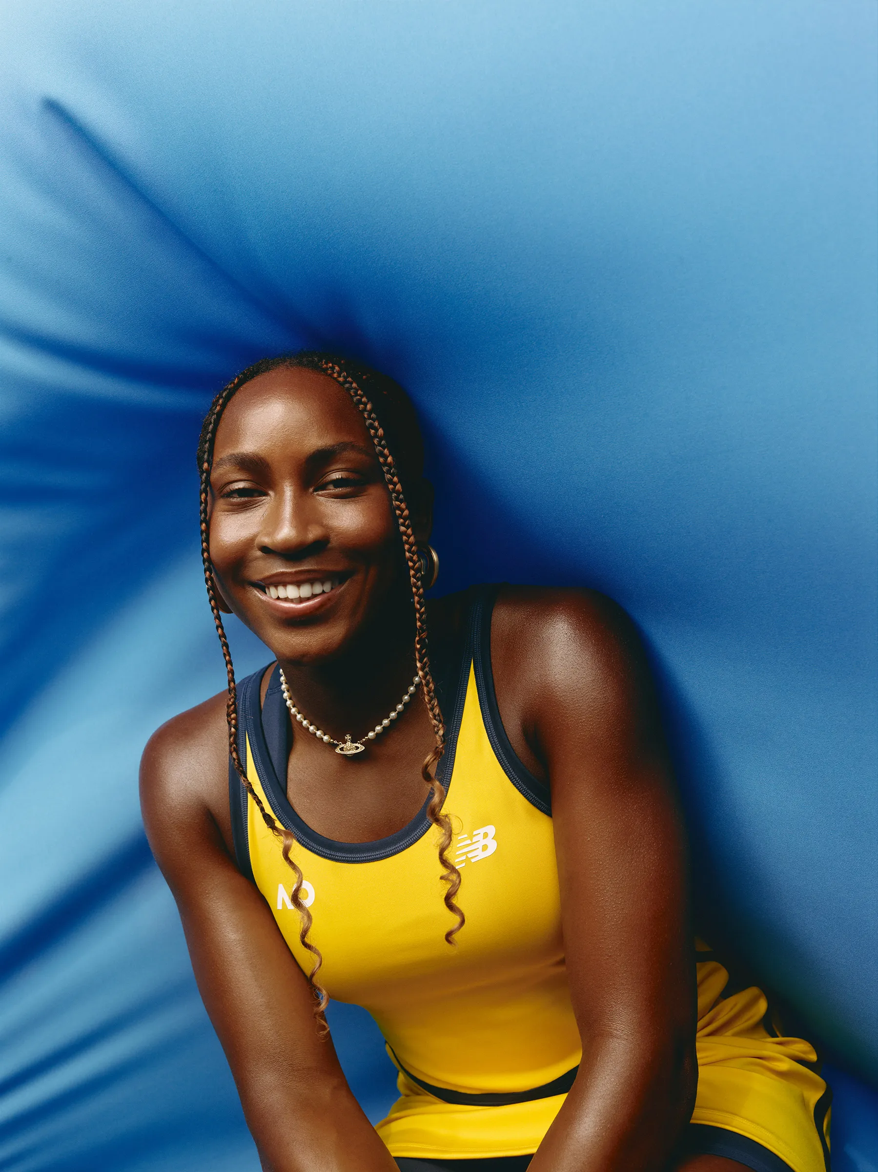 Portrait of a smiling African-American woman with braided hair, wearing a yellow tennis dress with the New Balance logo against a blue background.