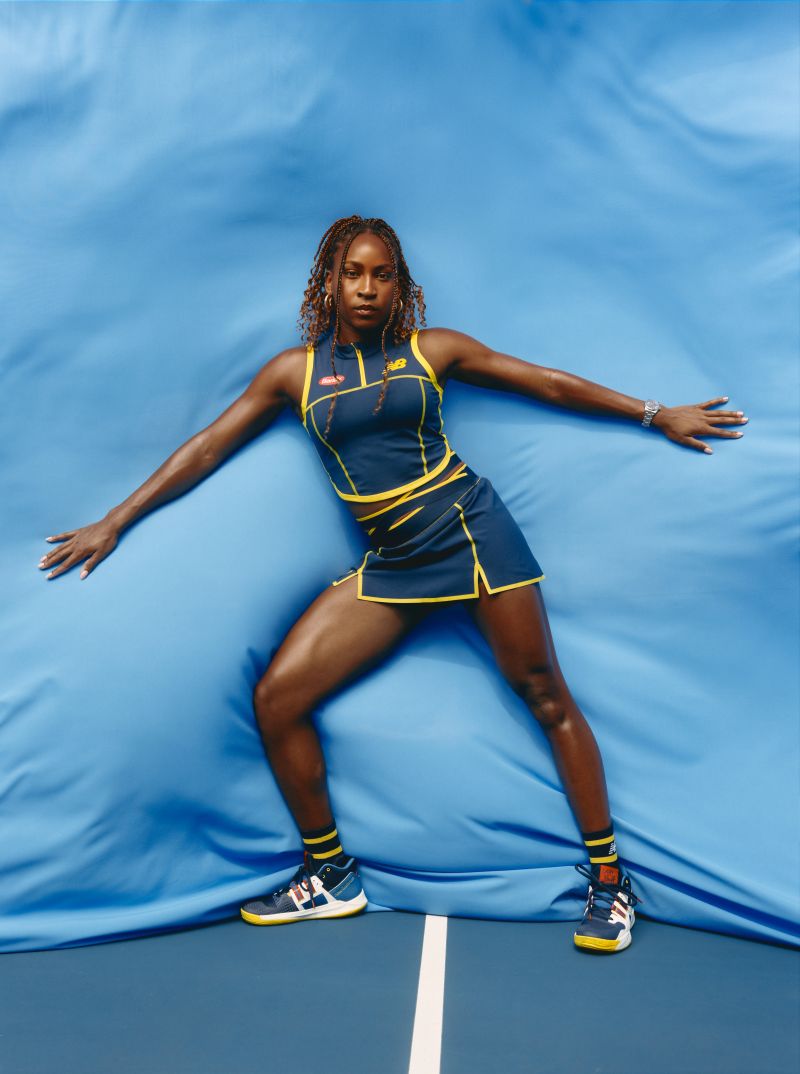 Professional portrait of a Black female athlete in a navy tennis outfit with yellow trim, posing against a draped light-blue background.