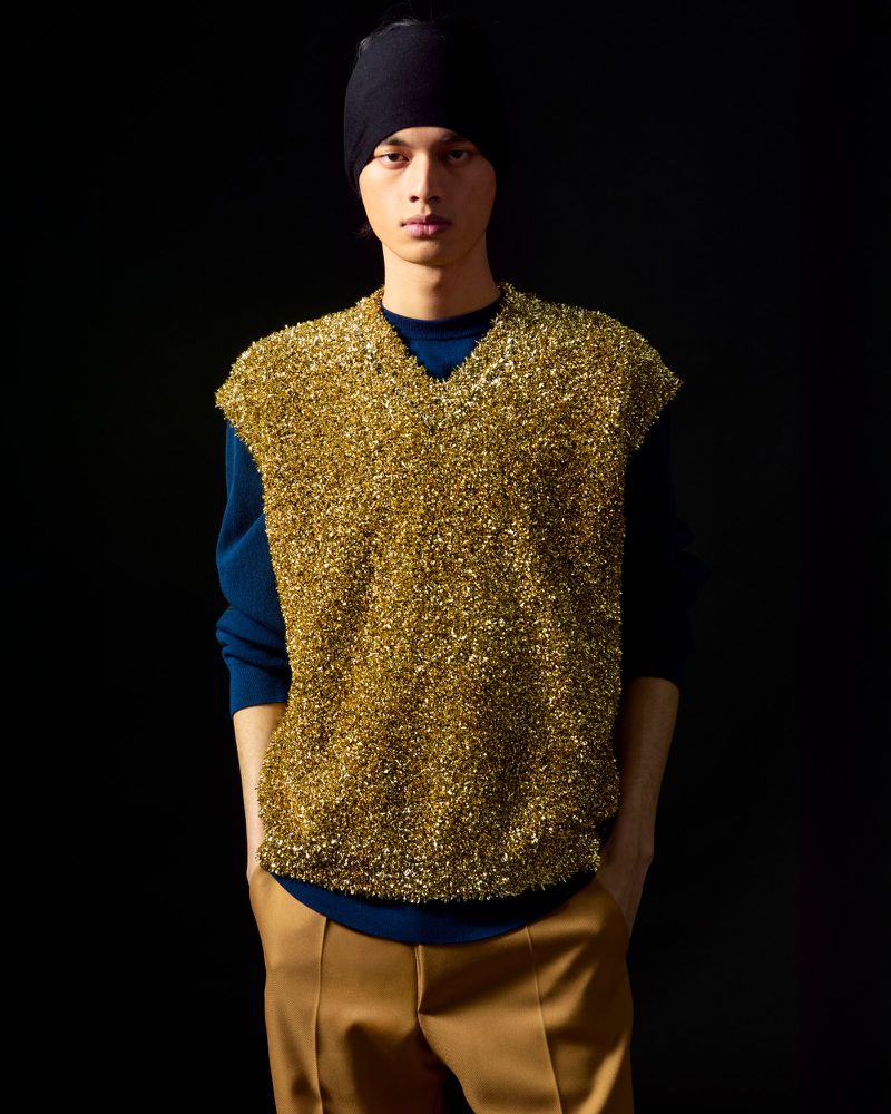 Male fashion model poses in a studio wearing a sparkling gold vest, a blue sweater, khaki pants, and a black beanie hat.