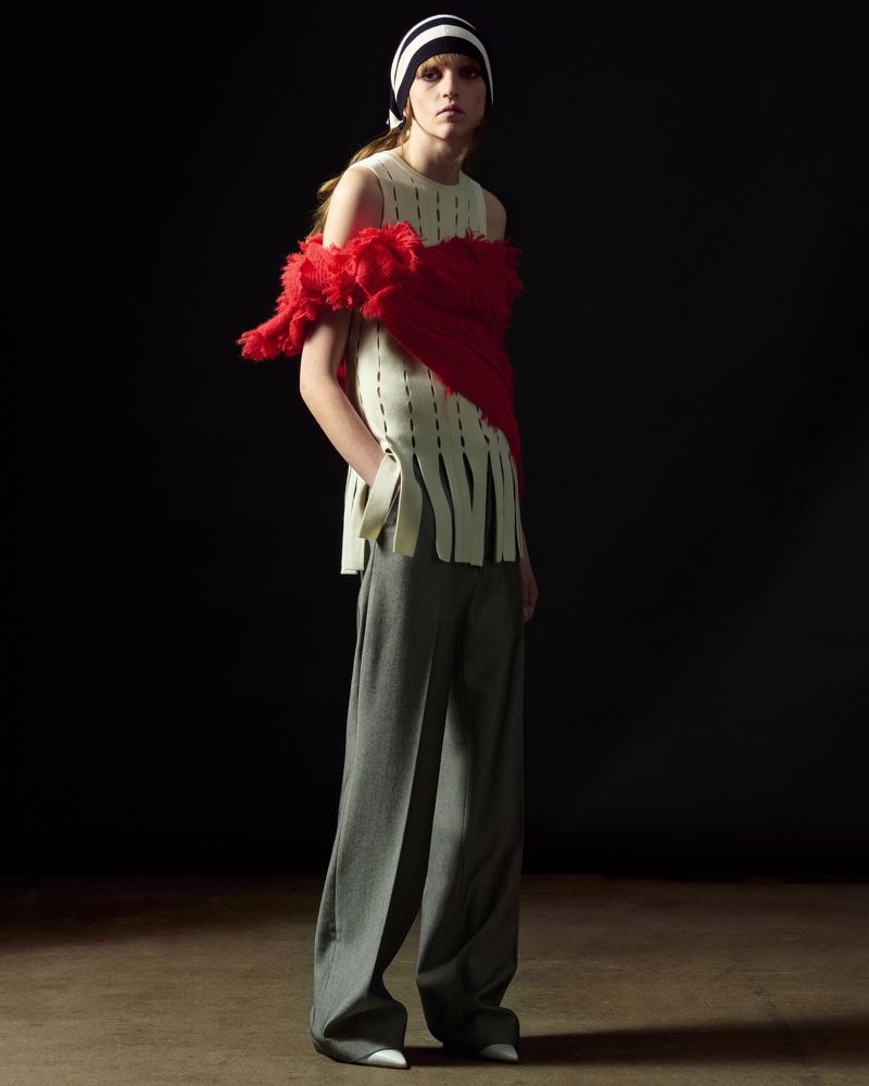 Model in striped cap, cream knit top with red feather wrap, and grey trousers against a dark background