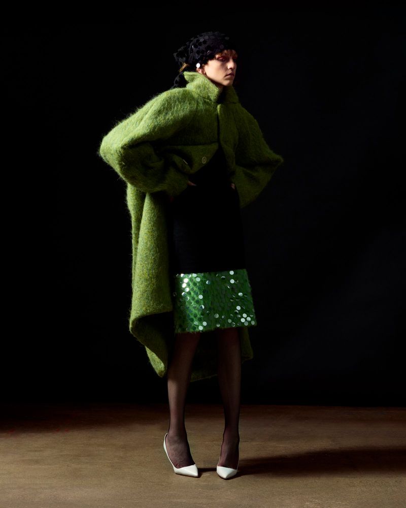 Fashion shot of an adult woman posing in a green coat and sequined skirt in a photo studio against a dark background.