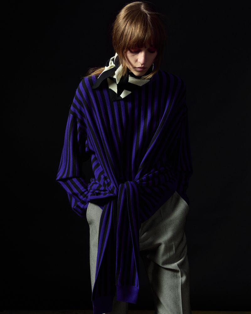 Fashionable woman in striped purple top and gray pants poses against a dark background, wearing a scarf, in studio lighting.