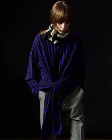 Fashionable woman in striped purple top and gray pants poses against a dark background, wearing a scarf, in studio lighting.