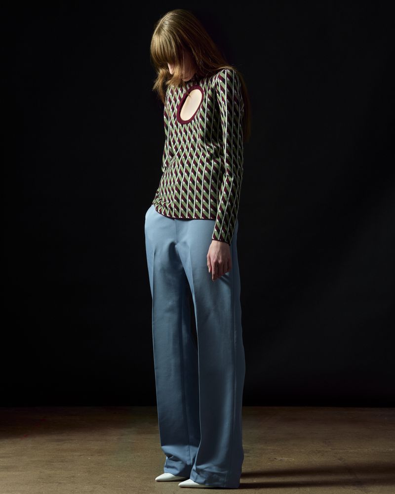 Full-length studio shot of a slender adult woman wearing a patterned top and blue trousers looking down against a black background.
