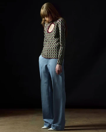 Full-length studio shot of a slender adult woman wearing a patterned top and blue trousers looking down against a black background.