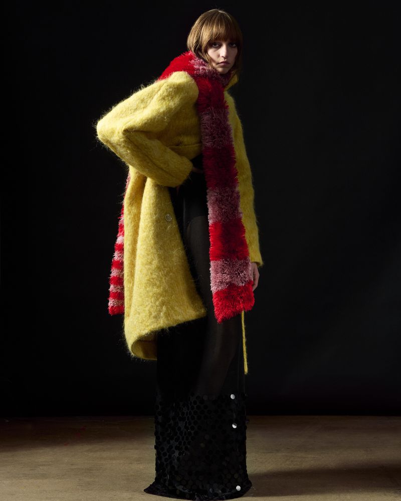 Model wearing a fuzzy yellow coat and a long red scarf against a dark background