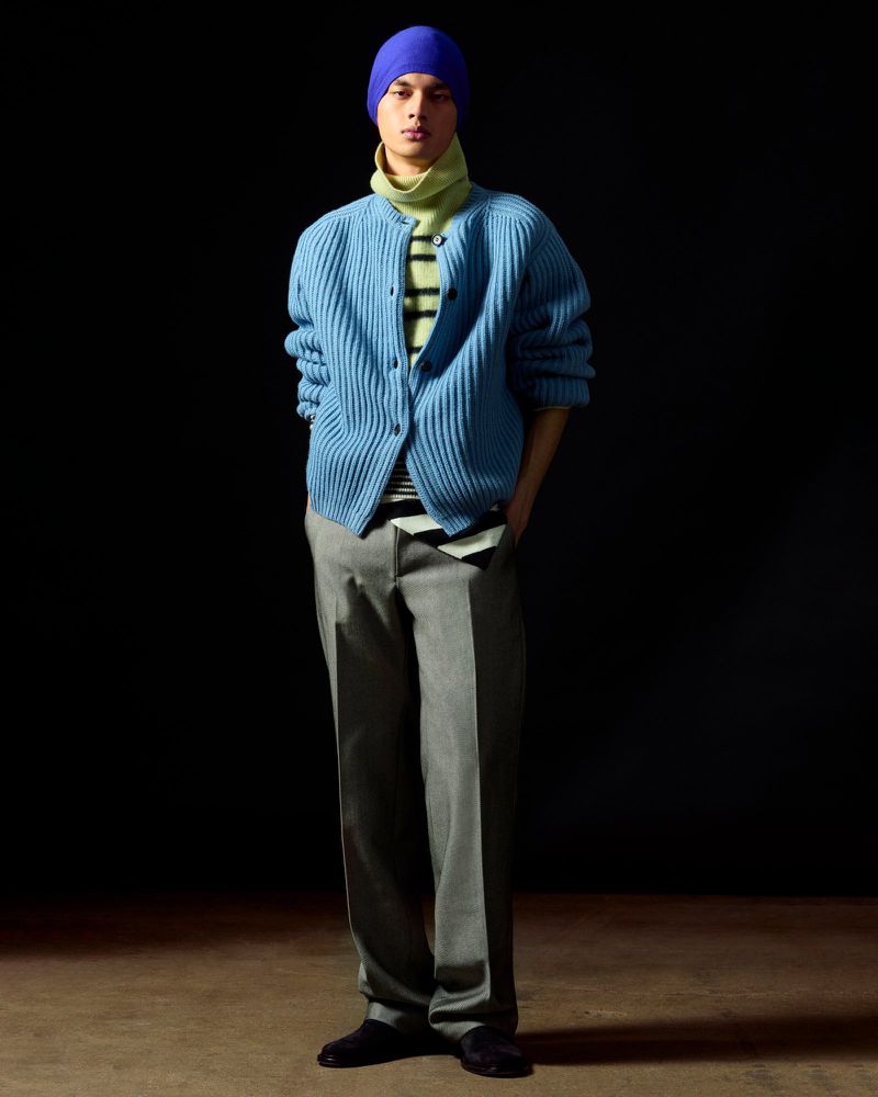 Full-shot of a male model in a blue cardigan, gray trousers, and blue beanie posing against a dark background in a studio setting.