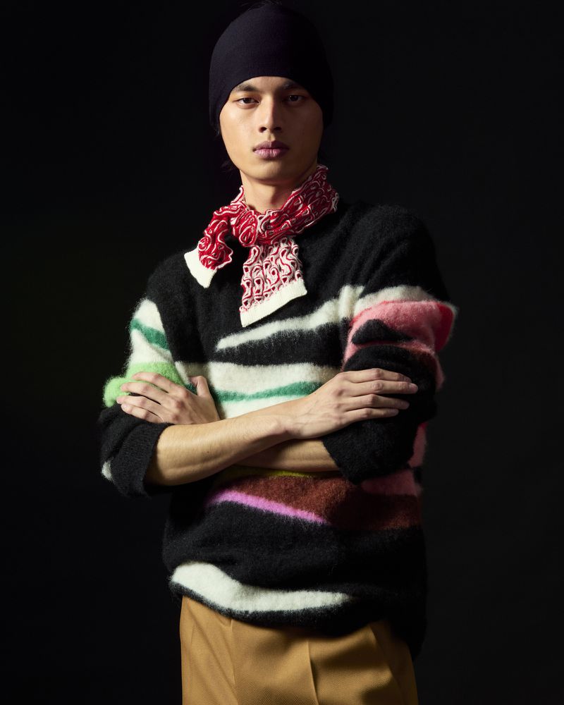 Model in a black beanie and striped sweater with a red patterned scarf against a dark background for Zankov FW25.