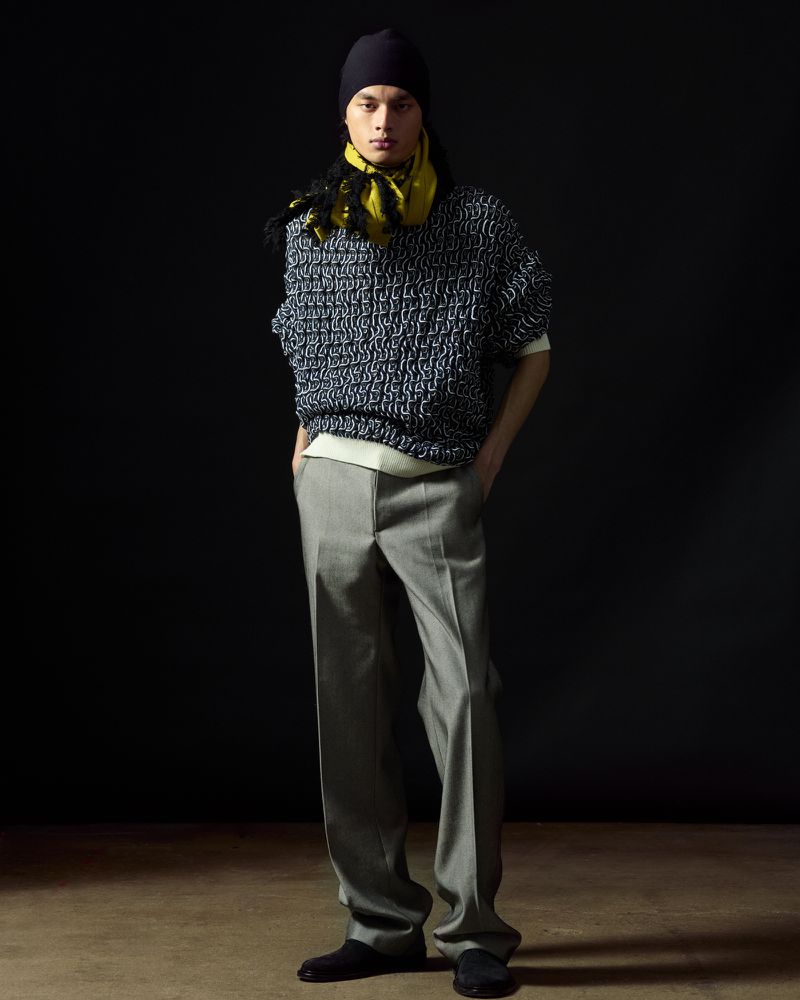 Male fashion model poses in oversized knit sweater, grey trousers, beanie and yellow scarf against a dark backdrop in a studio.