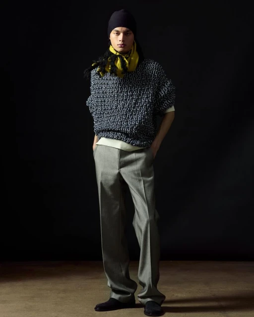 Male fashion model poses in oversized knit sweater, grey trousers, beanie and yellow scarf against a dark backdrop in a studio.