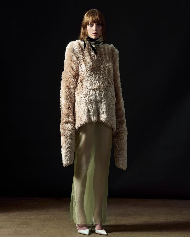 Fashion shot of a woman in a faux fur coat and sheer skirt, standing in front of a dark background in a photo studio with hardwood floors.