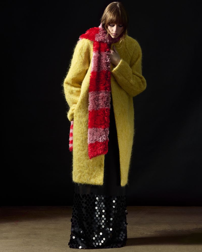 Full-length portrait of an adult woman in a yellow coat with a red scarf, posing in front of a dark background.