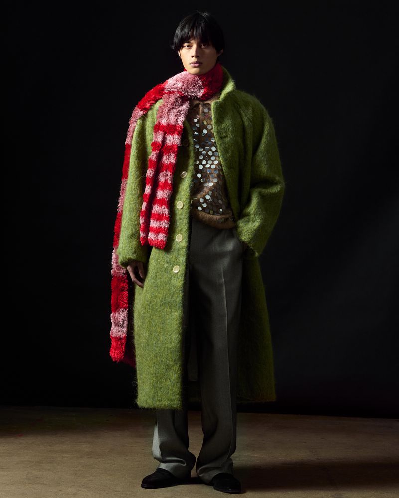 Model wearing a green textured coat and red patterned scarf standing against a dark backdrop for Zankov FW25.
