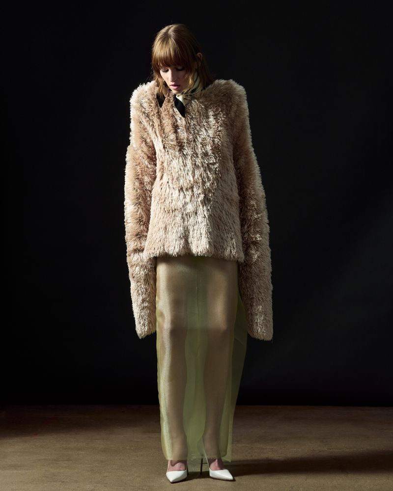 Fashion shot of a female adult posing in a beige faux-fur jacket and a sheer skirt against a dark background in a photo studio.