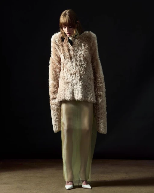 Fashion shot of a female adult posing in a beige faux-fur jacket and a sheer skirt against a dark background in a photo studio.