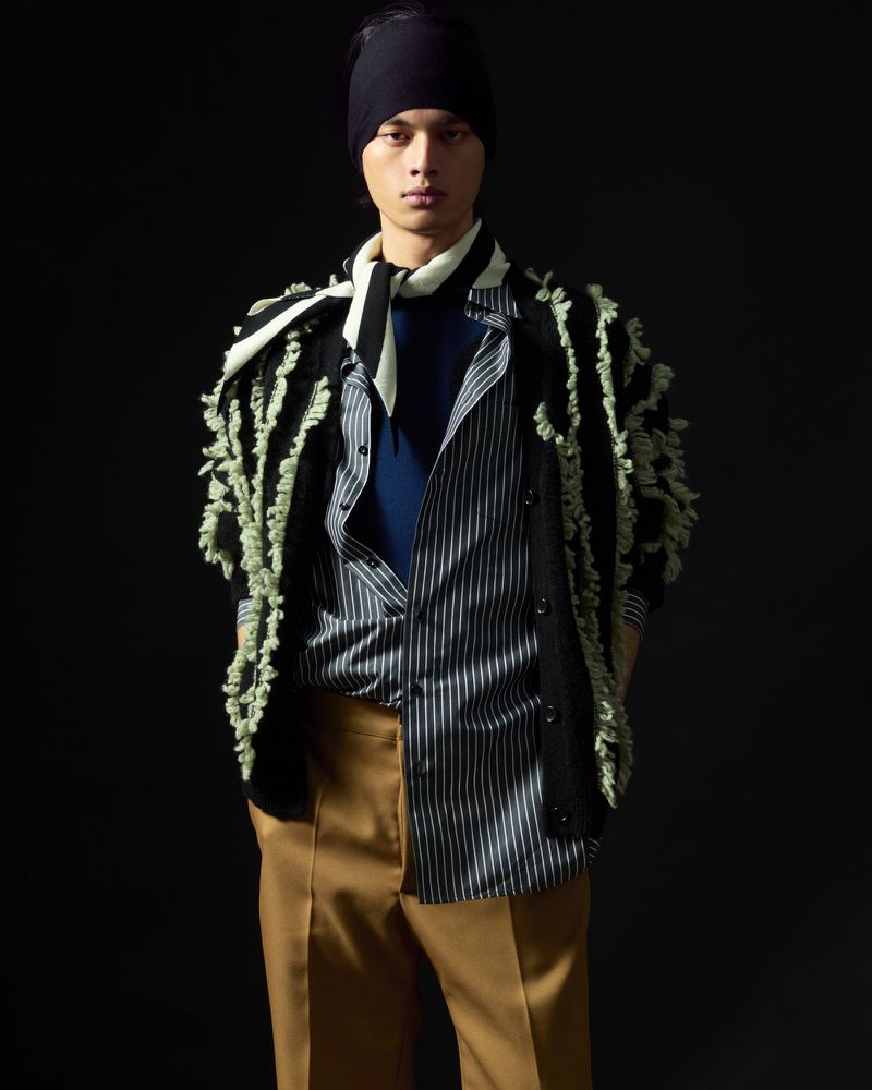 Portrait of an adult male wearing a striped shirt, cardigan, scarf, and head covering, posing against a black backdrop.