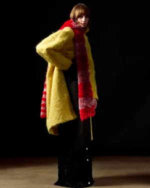 Model wearing a fuzzy yellow coat and a long red scarf against a dark background