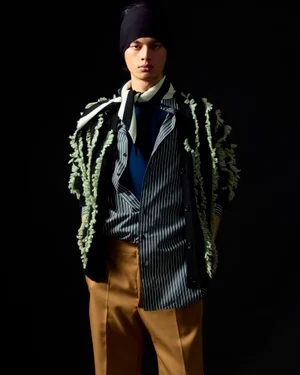 Model wearing a black cap, striped shirt, camel trousers, and textured cardigan against a plain black background