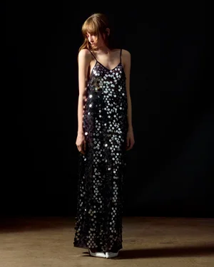 Model in a black sequin slip dress and white pointed heels standing against a dark background