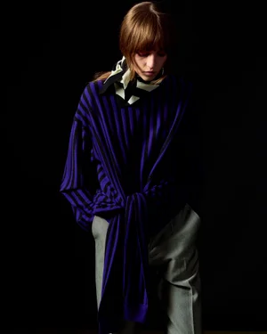 Model in a striped purple sweater and grey trousers against a black background for Zankov FW25.