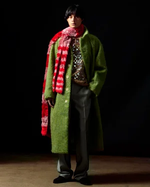Model wearing a green textured coat and red patterned scarf standing against a dark backdrop for Zankov FW25.