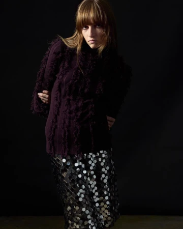 Model wearing a dark purple fringed sweater and metallic sequin skirt against a solid black studio background.